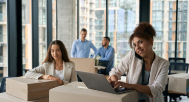 Joyful company worker working on laptop during phone conversation while her colleagues packing cardboard boxes